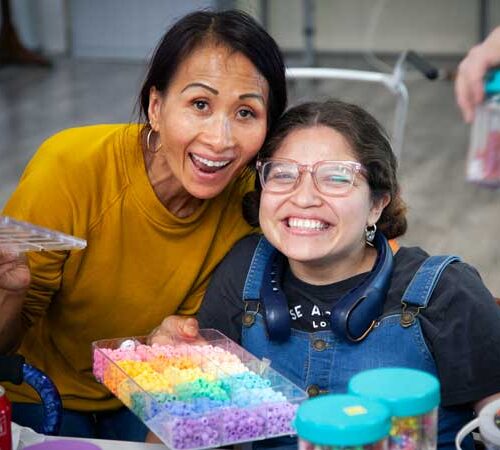 A petite south-east Asian woman dressed in a yellow sweater with dark hair and hoop earings displays a box of sorted beads next to a seated latina woman wearing pink glasses and overalls.