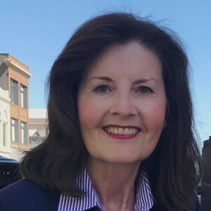 Headshot image of Director Louise Nathe, a White woman with dark brown hair wearing a blue-striped collared shirt under a blue jacket.
