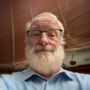 Portrait of Kevin Hanratty, a White man with mostly white hair and a bushy white beard. He is wearing browline glasses and a blue button-up shirt.