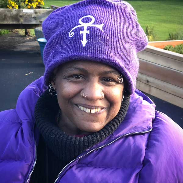 Profile image of Monique Harris, a smiling Black woman wearing a purple jacket and knit beanie over a dark turtlenecck. She has several peircings on her eyebrows, nose and ears.