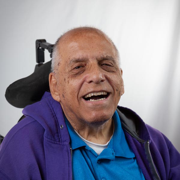 Profile image of John, a tan-skinned White man with cropped gray hair wearing a blue polo shirt and purple hoodie. John is smiling and seated in a power chair in front of a white backdrop.
