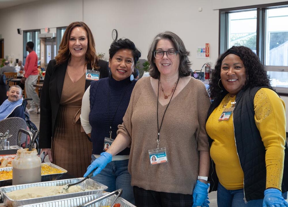 Ability Now Bay Area staff posed behind a row of large food containers prepared to serve at an event.